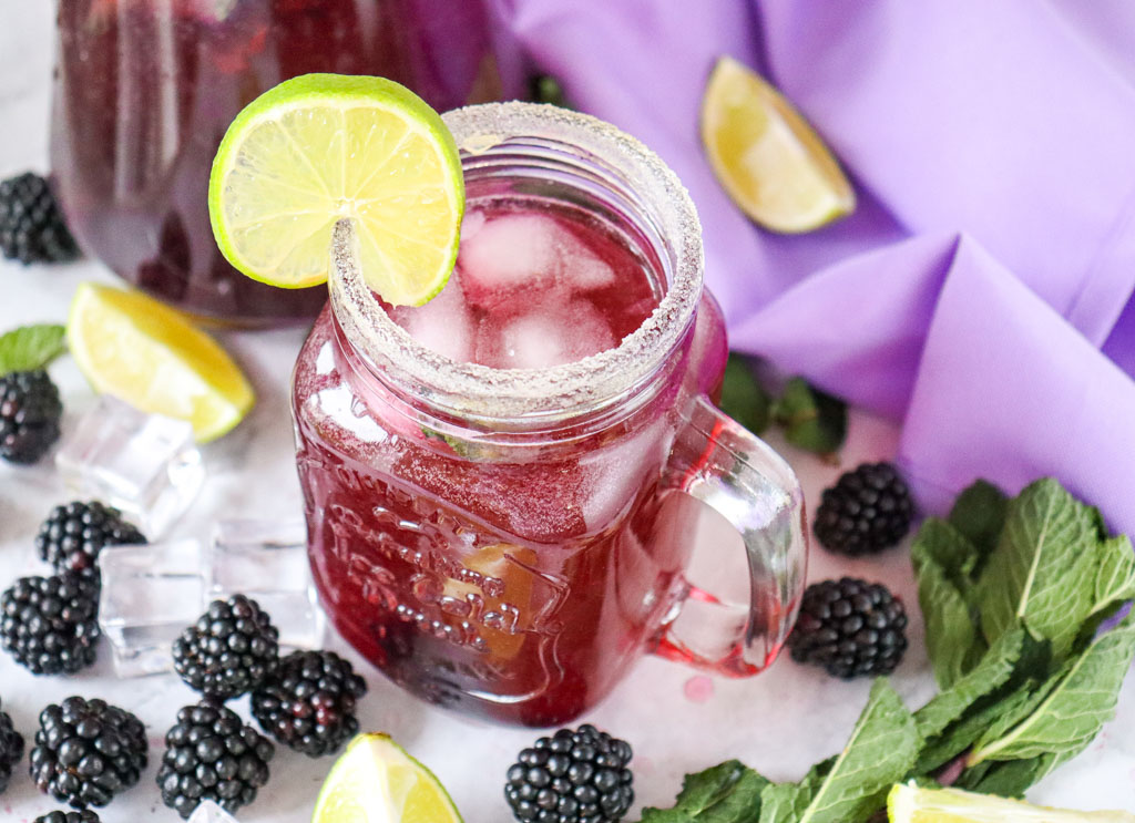 A tall glass of blackberry lemon mocktail with sparkling fizz, fresh mint, lime slices, and whole blackberries for garnish.