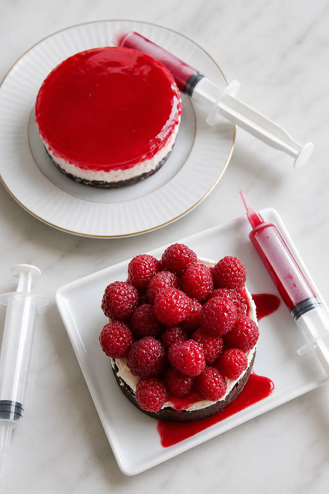 This Bloody Halloween Cheesecake has a chocolate crust, creamy no-bake filling, fresh raspberries, and raspberry “blood” sauce with food-safe syringes.