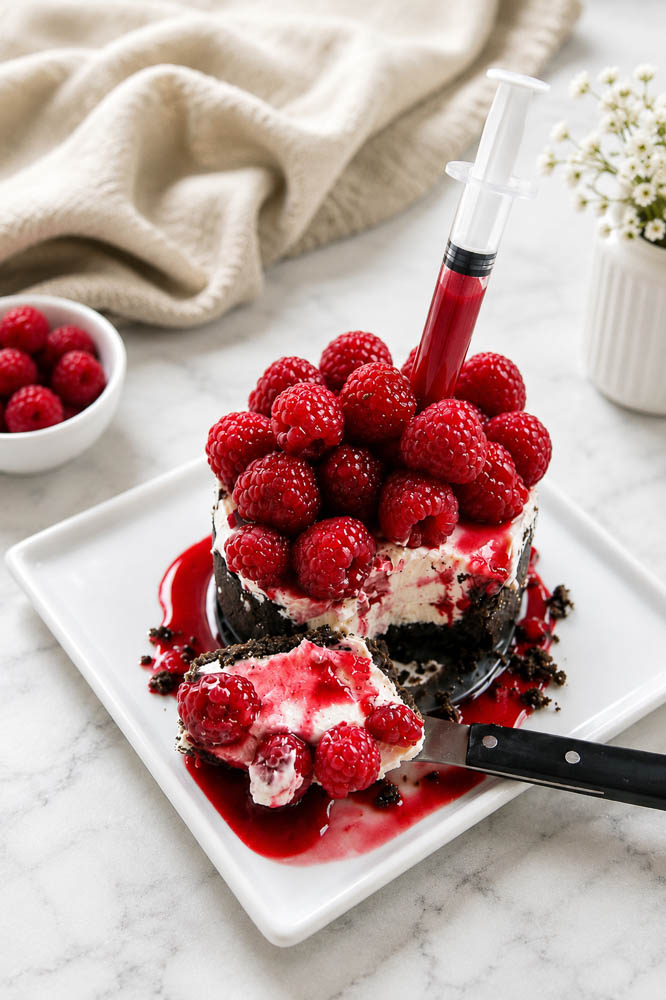 This Bloody Halloween Cheesecake has a chocolate crust, creamy no-bake filling, fresh raspberries, and raspberry “blood” sauce with food-safe syringes.